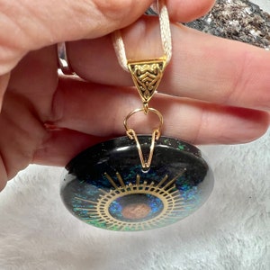May include: A round black pendant featuring a central gold-colored sunburst design with a brown circular accent. The pendant is embedded with iridescent green and blue flecks, suspended from a white ribbon by a gold-toned triangular metal bail.