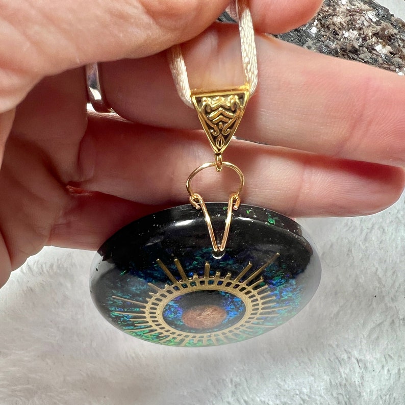 May include: A round black pendant featuring a central gold-colored sunburst design with a brown circular accent. The pendant is embedded with iridescent green and blue flecks, suspended from a white ribbon by a gold-toned triangular metal bail.