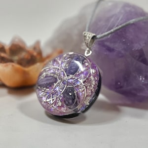 Op de afbeelding: Een ronde hangerketting met een zilveren beugel en grijs koord. De hanger is een mix van paars en zilver, met een zwarte basis. De hanger heeft een gelaagd ontwerp met een geometrisch patroon. De ketting is waarschijnlijk een sieraad.