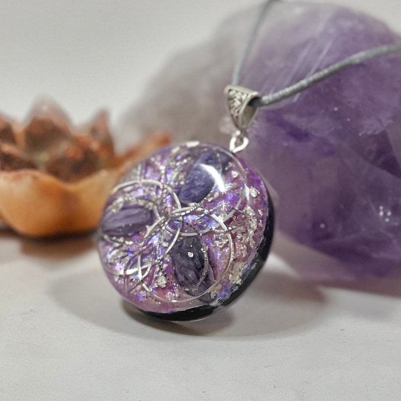 Op de afbeelding: Een ronde hangerketting met een zilveren beugel en grijs koord. De hanger is een mix van paars en zilver, met een zwarte basis. De hanger heeft een gelaagd ontwerp met een geometrisch patroon. De ketting is waarschijnlijk een sieraad.