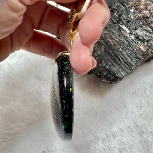 May include: A round, black pendant with embedded iridescent glitter and gold-colored flecks, featuring a gold-toned metal bail at the top. The pendant is suspended from a light-colored cord and displays a smooth, glossy, and slightly domed surface.
