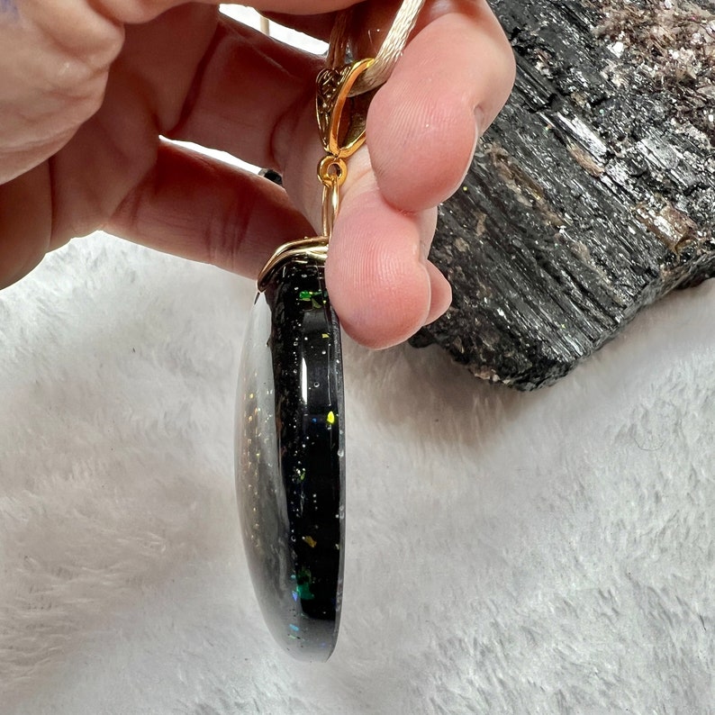 May include: A round, black pendant with embedded iridescent glitter and gold-colored flecks, featuring a gold-toned metal bail at the top. The pendant is suspended from a light-colored cord and displays a smooth, glossy, and slightly domed surface.