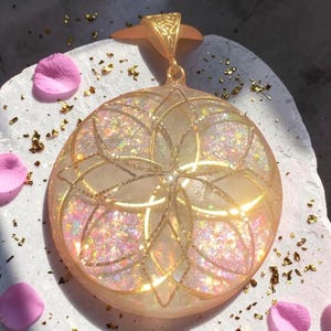Könnte beinhalten: Ein runder Anhänger mit einer durchscheinenden, pfirsichfarbenen Harzbasis, die mit irisierendem Glitzer und Blattgoldflocken durchsetzt ist. Die Oberfläche zeigt ein goldenes, geometrisches Blumenmuster mit geschwungenen Blütenblättern.