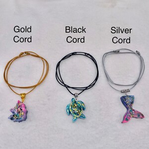 May include: Three necklaces with resin pendants: a pink and blue unicorn on a gold cord, a blue and green sea turtle on a black cord, and a blue and pink mermaid tail on a silver cord. Text labels read Gold Cord, Black Cord, and Silver Cord.