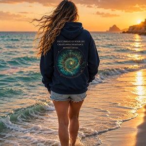 May include: A dark navy blue hooded sweatshirt featuring a circular graphic on the back with a swirling galaxy design in shades of teal, gold, and green. The text above the graphic reads: THE CURRENT OF YOUR LIFE CREATES WHAT RESISTANCE NEVER COULD.
