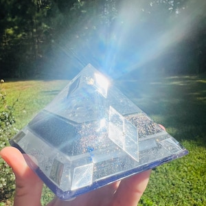 May include: A clear, pyramid-shaped resin art piece with embedded glitter and geometric accents. The pyramid is held up against a backdrop of green grass and trees, with sunlight shining through the pyramid.