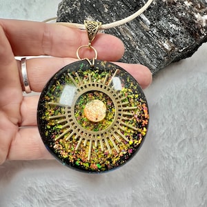 May include: A gold pendant necklace with a round, black resin pendant featuring a gold sunburst design with iridescent glitter embedded in the resin.