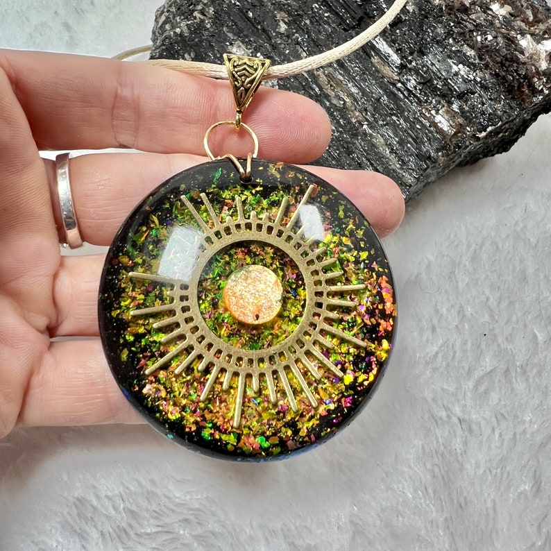 May include: A round resin pendant featuring a black base embedded with iridescent green and gold flakes. A gold-toned metal sunburst charm is centered on the surface, surrounding a small, textured orange circular inlay. The pendant hangs from a cord.