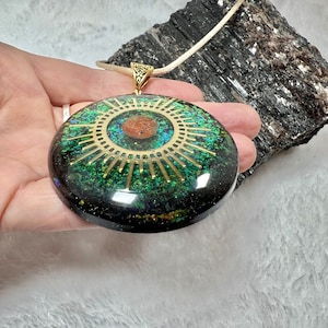 May include: A circular pendant featuring a dark resin base embedded with green and blue iridescent flakes. A gold-toned sunburst emblem with a central copper-colored circle is set into the surface. The pendant hangs from a light-colored cord.