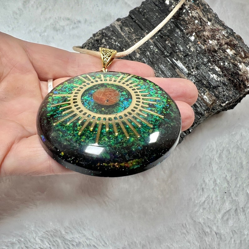 May include: A circular pendant featuring a dark resin base embedded with green and blue iridescent flakes. A gold-toned sunburst emblem with a central copper-colored circle is set into the surface. The pendant hangs from a light-colored cord.