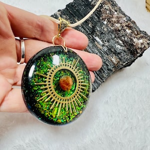 May include: A round resin pendant featuring a black and green glitter background with a gold-toned sunburst metal charm and a small brown spherical bead at the center. The pendant is attached to a light-colored cord with a gold-toned metal bail.