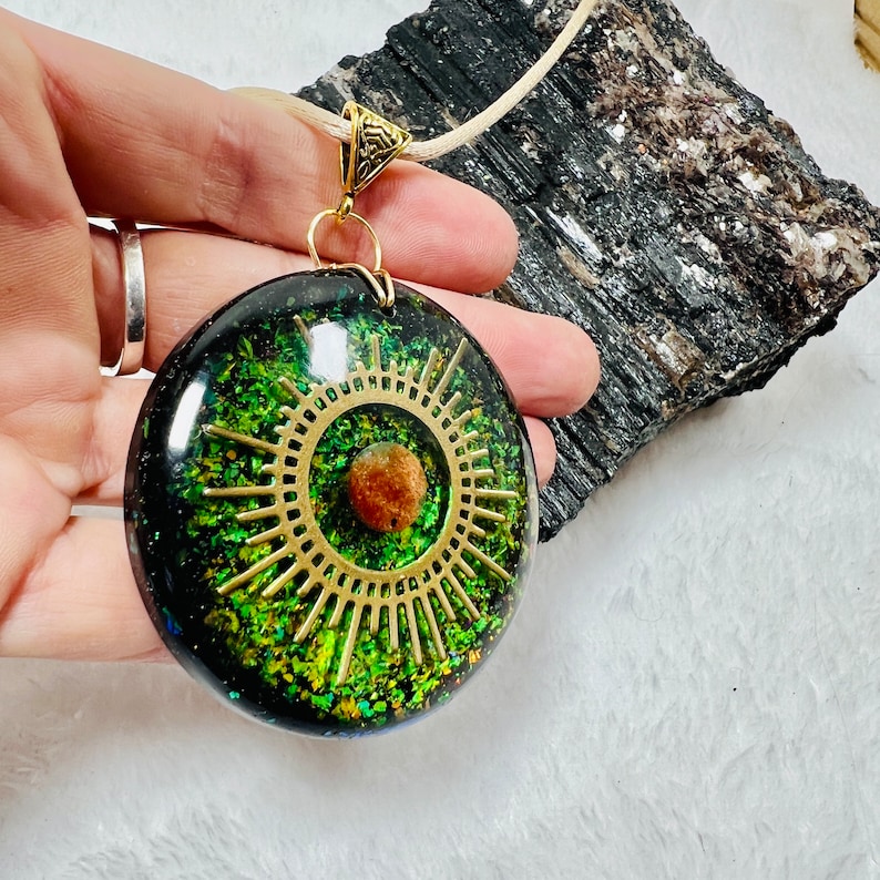 May include: A round resin pendant featuring a black and green glitter background with a gold-toned sunburst metal charm and a small brown spherical bead at the center. The pendant is attached to a light-colored cord with a gold-toned metal bail.