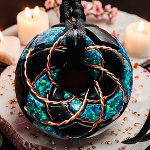 Può includere: Un pendente sferico con base nera, intarsiato con frammenti iridescenti blu-verdi e dettagli in filo di rame. Il pendente è sospeso a un cordino nero. Lo sfondo include candele accese e accenti floreali.
