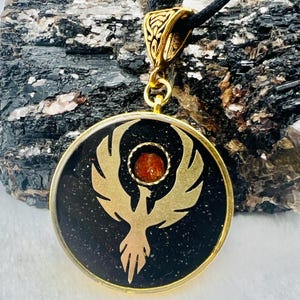 Może przedstawiać: Złoty wisiorek ze złotym wzorem feniksa na czarnym tle. Okrągły wisiorek ma mały, okrągły, pomarańczowy kamień pośrodku. Wisi na czarnym sznurku.