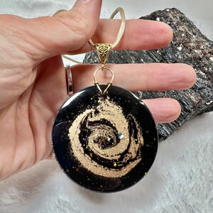 May include: A round pendant featuring a black base with a swirling gold glitter pattern that resembles a galaxy. The pendant is attached to a gold-toned metal bail and hangs from a light-colored cord. The surface is smooth, glossy, and reflective.