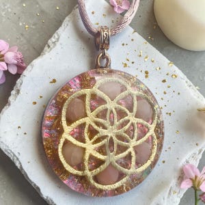 Op de afbeelding: Een ronde hanger met een goudkleurig geometrisch levensbloemmotief ingebed in heldere hars. De hars bevat roze getinte elementen, bladgoudvlokken en kleine gladde roze stenen, hangend aan een lichtroze zijden koord.