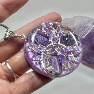 Op de afbeelding: Een ronde, paarse hangerketting met een zilverkleurige beugel en koord. De hanger heeft een heldere harskoepel met ingebedde paarse stenen en zilverkleurig filigraan. De achtergrond is lichtpaars.