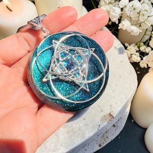 May include: A circular pendant featuring a deep teal resin base with embedded glitter. A silver-toned metal star-shaped frame with overlapping circular wire accents is set into the center. The pendant includes a silver-toned metal bail at the top.