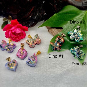 May include: A collection of resin pendants featuring various shapes including butterflies, a heart, a unicorn, and three dinosaurs labeled Dino #1, Dino #2, and Dino #3. Each pendant contains colorful glitter and metallic flakes with gold-toned bails.