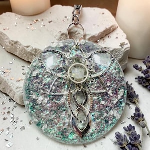 May include: A circular resin pendant featuring a mix of iridescent purple, green, and silver flakes embedded within the clear material. A silver-toned metal overlay in a floral and infinity knot design sits on top, centered with a small clear stone.