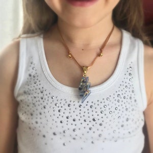 May include: A white tank top featuring a scattered pattern of silver rhinestones across the chest, paired with a necklace consisting of a brown cord and a lightning bolt pendant with a blue and gold glitter finish and a gold-toned metal bail.