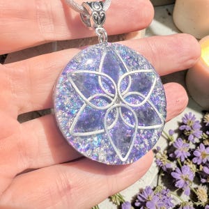 Puede incluir: Un colgante circular con un diseño de flor de metal plateado incrustado en una base de resina translúcida. La resina contiene fragmentos de piedra en tonos violetas y purpurina iridiscente que refleja la luz.