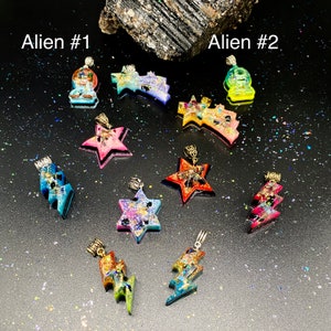 May include: A collection of ten resin pendants in various shapes including stars, lightning bolts, and shooting stars. The pieces feature vibrant colors, glitter, and metallic flakes. Two pendants are labeled Alien #1 and Alien #2.