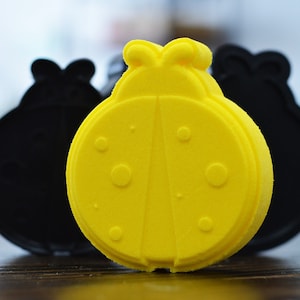 May include: Three ladybug-shaped bath bombs, one yellow and two black. The yellow bath bomb has black spots and a raised design.