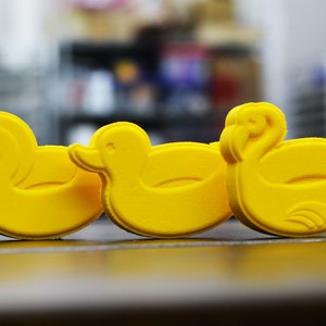 May include: Three yellow rubber duck figurines. The ducks are in a row, facing the right side of the image. The duck on the left has a curved beak and a rounded head. The duck in the middle has a flat beak and a rounded head. The duck on the right has a curved beak and a rounded head.