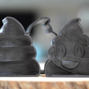 Puede incluir: Dos cortadores de galletas impresos en 3D en negro con forma de pila de caca. Un cortador tiene una cara sonriente con ojos y boca.