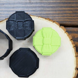 Peut inclure: Trois boîtes imprimées en 3D, deux noires et une verte. La boîte verte a un nœud sur le dessus. Les boîtes sont sur une surface en bois.