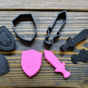 May include: A set of black and pink plastic cookie cutters shaped like swords and shields. The shields feature a castle design.