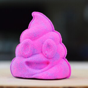 Puede incluir: Una bomba de ba&ntilde;o rosa y morada con forma de emoji de caca sonriente.