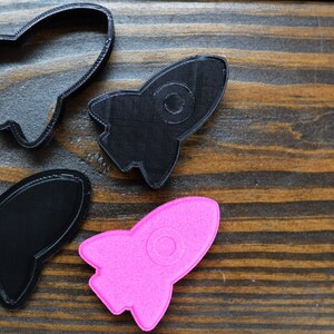 May include: Three rocket-shaped cookie cutters, two are black and one is pink. The cookie cutters are on a wooden surface.
