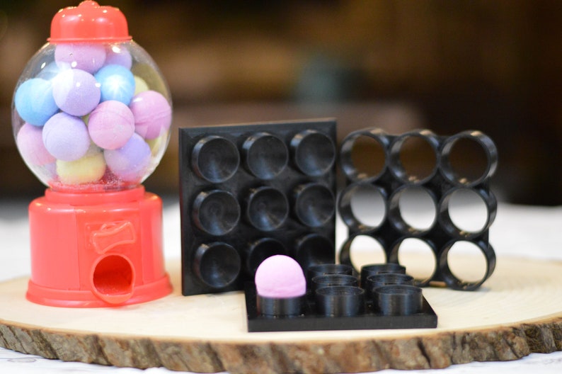 May include: A red gumball machine filled with colorful gumballs sits next to three black silicone fidget toys. The fidget toys have different shapes and sizes, including a grid of circles, a grid of squares, and a grid of half-circles.