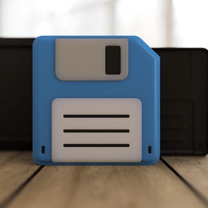 May include: A blue 3.5-inch floppy disk with a gray label and a white label with three horizontal lines. Two black rectangular objects are behind the floppy disk, all resting on a wooden surface.