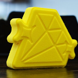 May include: A bright yellow, geometrically shaped, 3D printed object with a textured surface. The object is shaped like a diamond with a star-shaped design on each side.