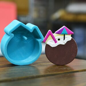 Peut inclure: Un moule en silicone bleu en forme de noix de coco avec un parasol rose et violet et une paille. Le moule est à côté d'une sucette en forme de noix de coco brune avec un parasol rose et violet et une paille.