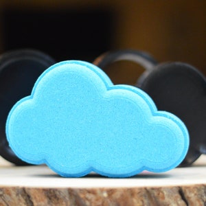 May include: A sky blue cloud-shaped bath bomb with a textured finish, set before two black cloud-shaped moulds. The objects are presented on a wooden surface, offering a contrast of colours and textures.
