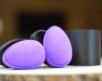 Puede incluir: Dos bombas de baño moradas en forma de huevo con una superficie texturizada, junto a un molde negro. Las bombas de baño están sobre una superficie de madera, con un fondo borroso. Las bombas de baño son de un tono morado vibrante.