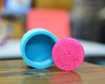 Op de afbeelding: Een blauwe siliconen mal met een roze siliconen deksel. De mal is rond en heeft een glad oppervlak. Het deksel heeft een getextureerd oppervlak.