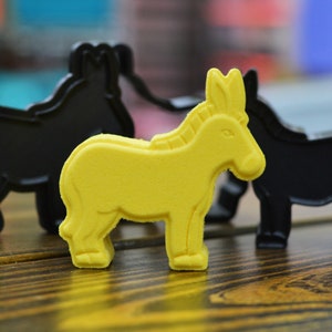 May include: Three toy animal figurines, two black and one yellow. The yellow figurine is a donkey, and the two black figurines are horses. The figurines are on a wooden surface.