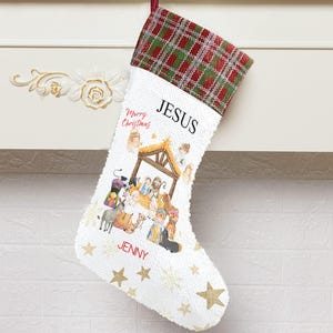May include: White Christmas stocking with a red and green plaid cuff. The stocking features the word "JESUS" and a nativity scene illustration. Gold stars and the name "JENNY" are also visible.
