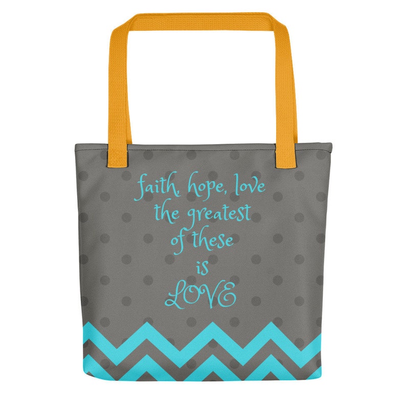K&ouml;nnte beinhalten: Eine graue und wei&szlig;e Einkaufstasche mit einem Zickzackmuster am Boden. Die Tasche hat gelbe Henkel und tr&auml;gt den Text "faith hope, love the greatest of these is LOVE" in Blau.