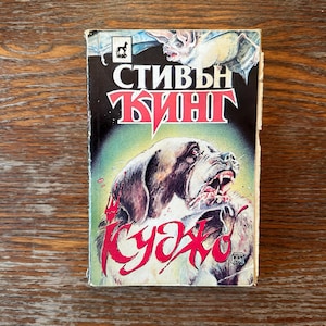 May include: A vintage paperback book with a cover featuring a snarling dog with blood around its mouth. The title "Cujo" is in red, with the author's name in Cyrillic script above. A bat is in the background.