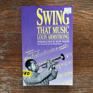 May include: A vintage book titled "Swing That Music" by Louis Armstrong. The cover is purple with yellow text and features a black and white image of Armstrong playing the trumpet. The book has a price tag.