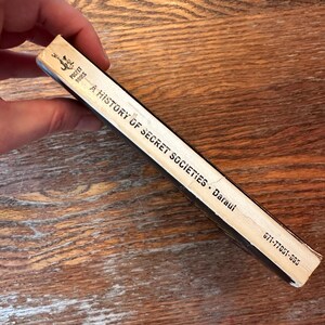 May include: A vintage paperback book titled "A History of Secret Societies" by Daraul. The book is a Pocket Books edition with a black spine and off-white pages. The book is held by a hand.