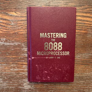 May include: A maroon book titled "Mastering the 8088 Microprocessor" by Lanny V. Dao. The text is in gold lettering. The book is on a wooden surface.