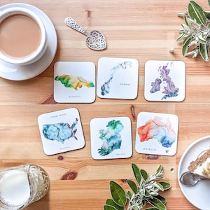 May include: A collection of square coasters featuring watercolor maps of Jamaica, Norway, the United Kingdom, the United States, Ireland, and Australia. Each coaster has the country's name printed below the map. A cup of tea, a decorative spoon, and greenery are also visible.