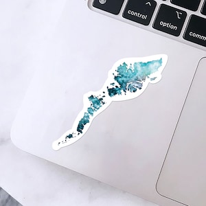 May include: A white sticker featuring a watercolor design of a landmass in shades of teal, blue, and gray. The sticker is placed on a white laptop, with the keyboard visible in the background.